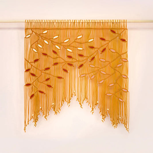 A golden yellow beaded fringe wall hanging with embroidered branch and leaf patterns, suspended from a light wooden rod against a plain off-white wall.