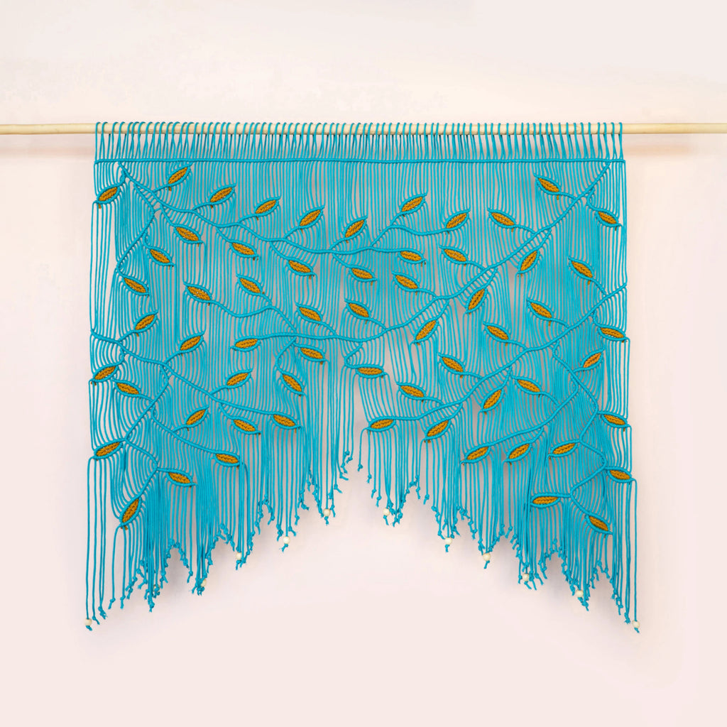 A turquoise blue fringed wall hanging or curtain with embroidered leaf-like details, suspended from a light wooden rod against a plain off-white wall.