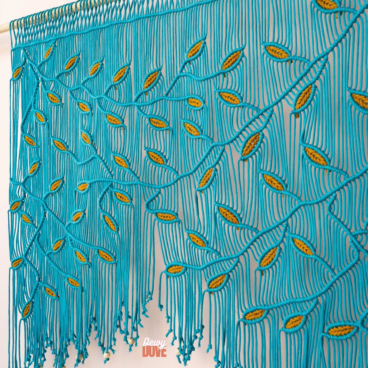 A close-up of a brown macrame wall hanging with intricate leaf and vine patterns and hanging tassels, shown against a light beige wall with a small 'Davy Cute' watermark at the bottom.