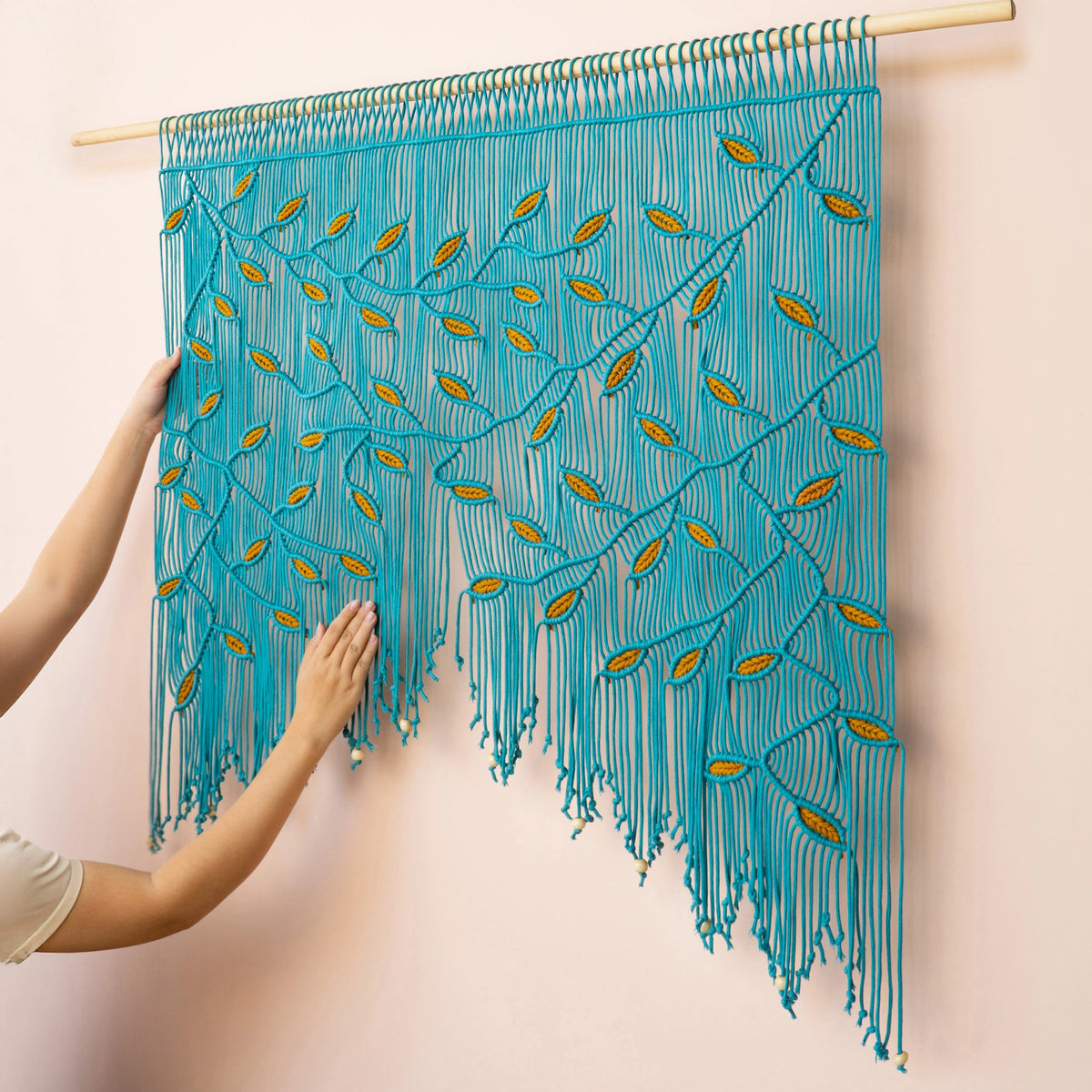 Wide horizontal macrame tapestry featuring a botanical leaf motif in blue and mustard hues, mounted on a solid wooden dowel.