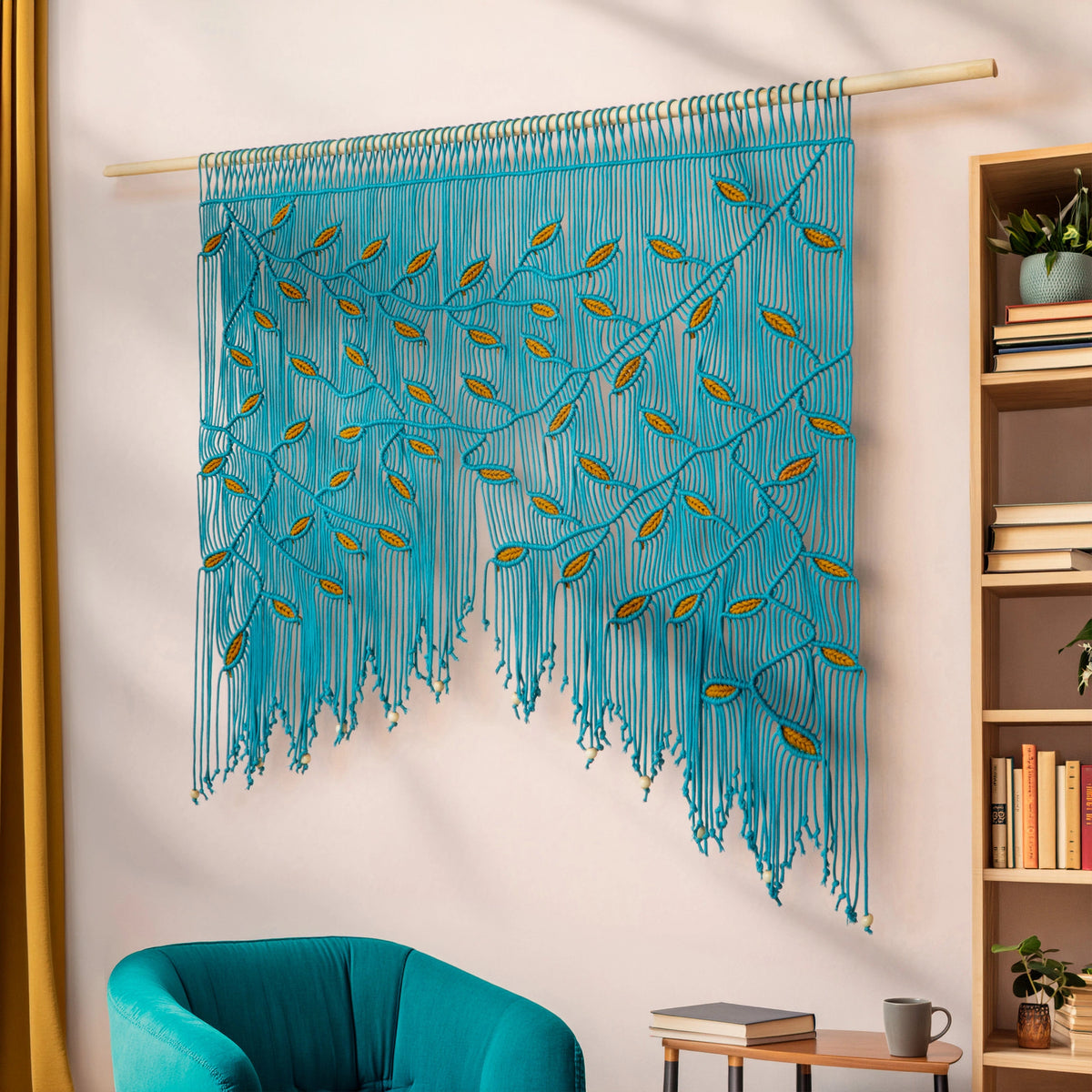 A turquoise blue bohemian wall hanging macrame wall tapestry with gold leaf accents, suspended from a wooden dowel in a styled room setting next to a teal armchair and a wooden bookshelf.