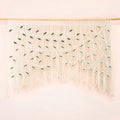 A bohemian-style macrame wall hanging with intricate woven leaf patterns and long fringe, suspended from a light wood rod against a soft pink background.