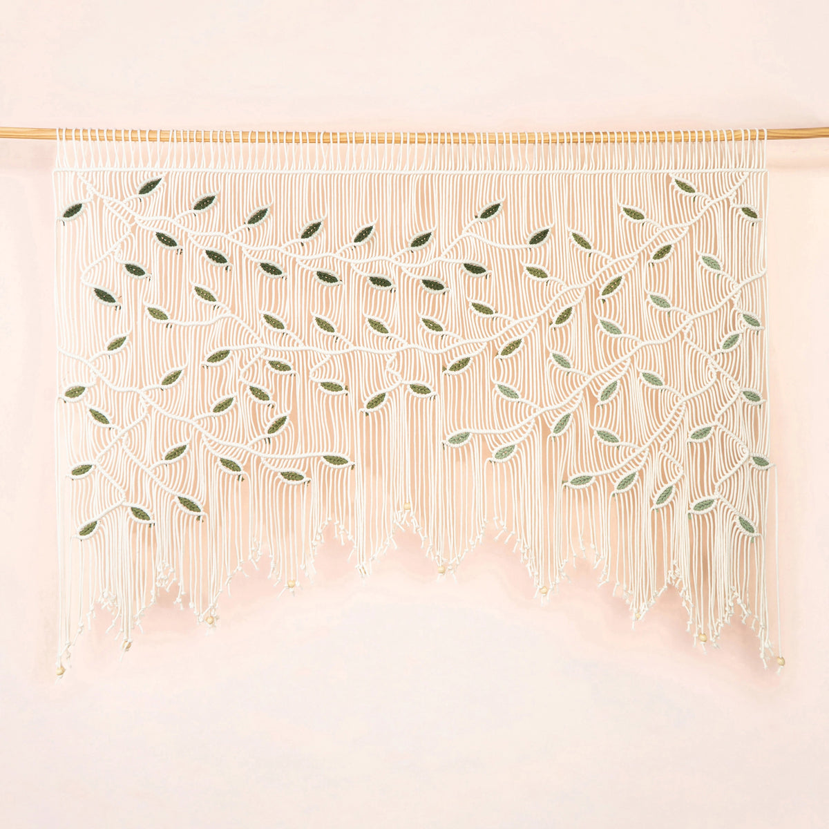 A bohemian-style macrame wall hanging with intricate woven leaf patterns and long fringe, suspended from a light wood rod against a soft pink background.