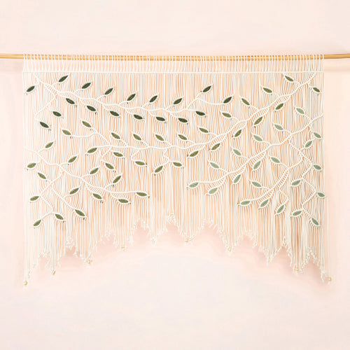 A bohemian-style macrame wall hanging with intricate woven leaf patterns and long fringe, suspended from a light wood rod against a soft pink background.