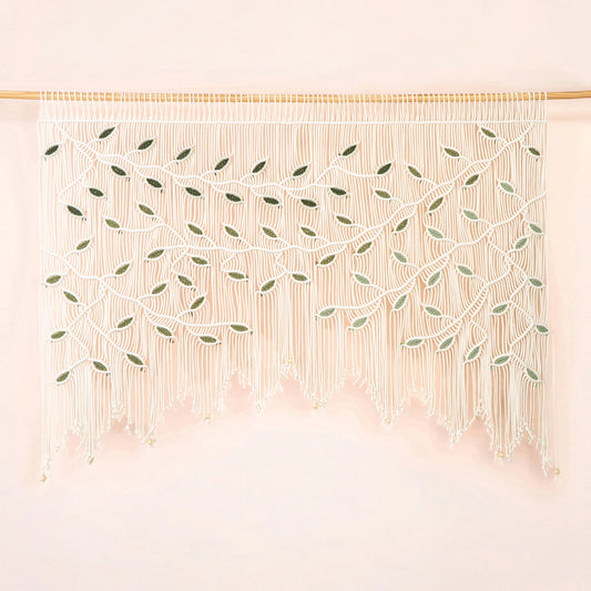 A bohemian-style macrame wall hanging with intricate woven leaf patterns and long fringe, suspended from a light wood rod against a soft pink background.