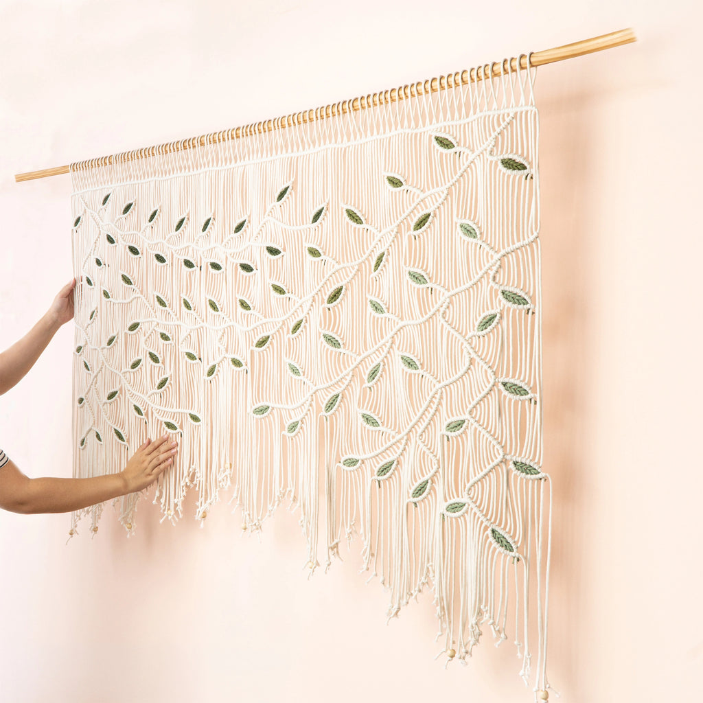 A large macrame wall hanging with a tree branch and leaf pattern, suspended from a wooden rod against a pale pink wall. A person's hands are visible on the left holding the hanging in place.