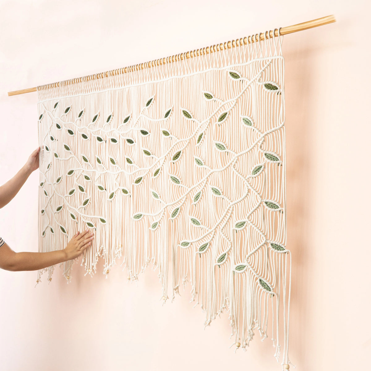 A large macrame wall hanging with a tree branch and leaf pattern, suspended from a wooden rod against a pale pink wall. A person's hands are visible on the left holding the hanging in place.