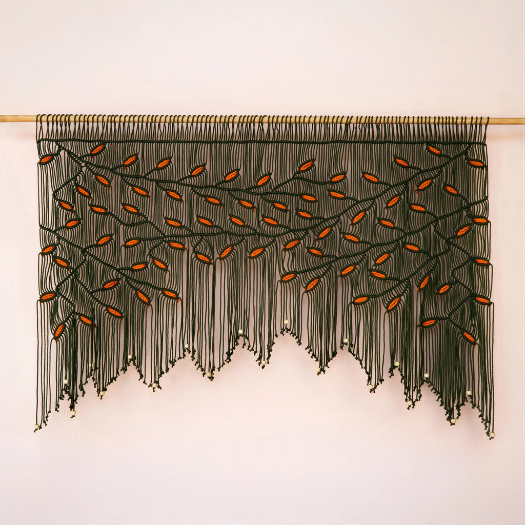 Large-scale dark green and orange leaf fiber tapestry installed on a light wood rod against a warm neutral accent wall.