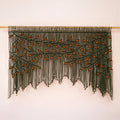 Large-scale dark green and orange leaf fiber tapestry installed on a light wood rod against a warm neutral accent wall.