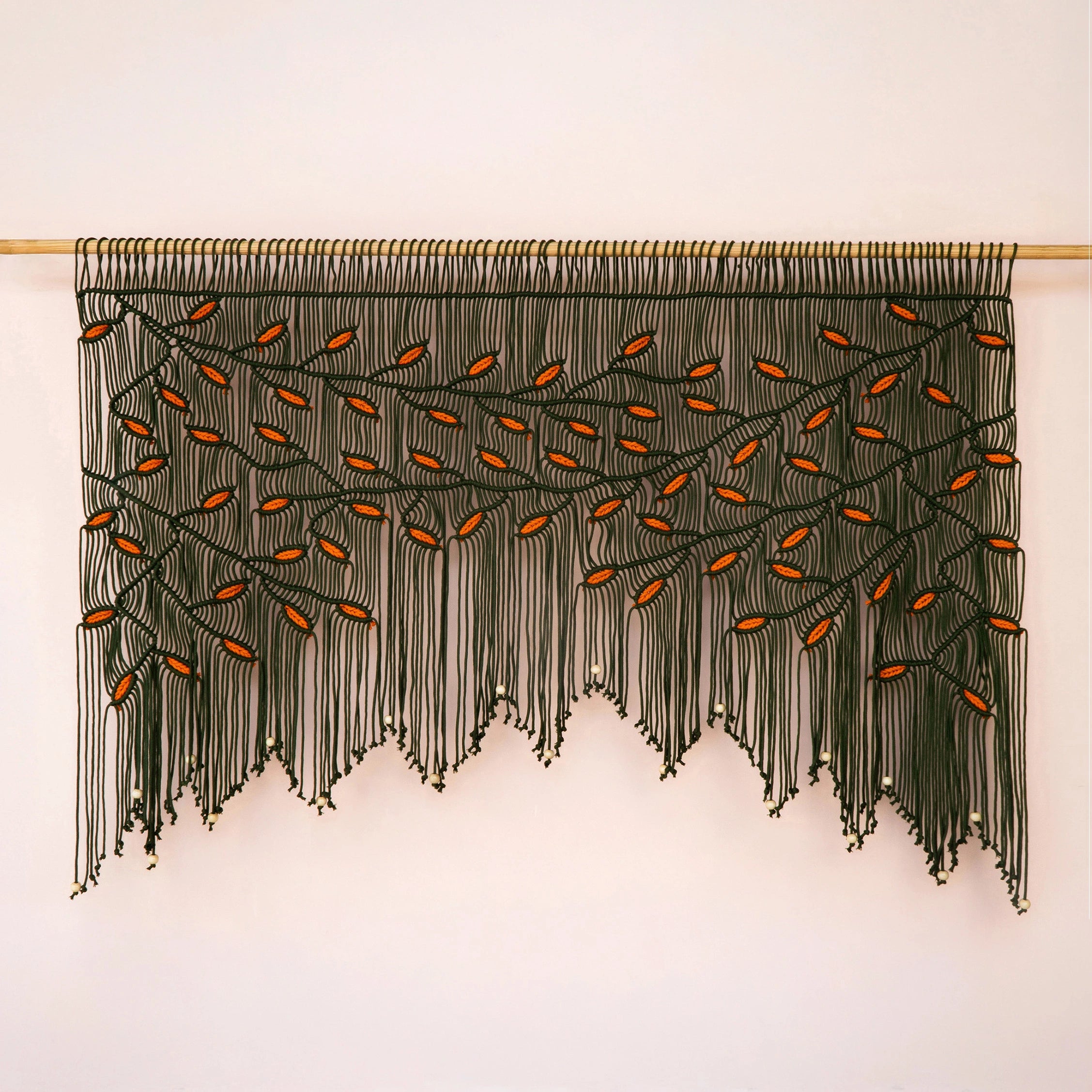 Large-scale dark green and orange leaf fiber tapestry installed on a light wood rod against a warm neutral accent wall.