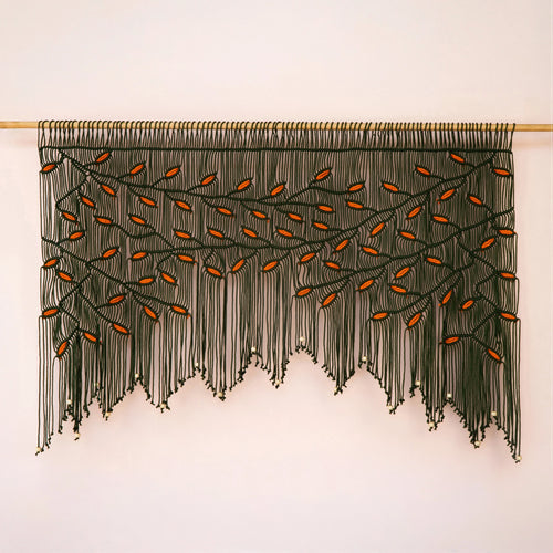 Large-scale dark green and orange leaf fiber tapestry installed on a light wood rod against a warm neutral accent wall.
