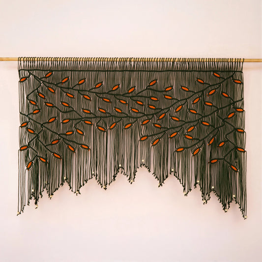 Large-scale dark green and orange leaf fiber tapestry installed on a light wood rod against a warm neutral accent wall.