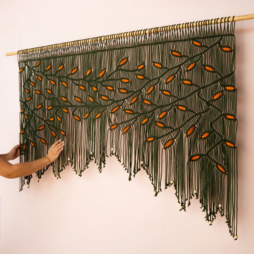 Wide horizontal macrame tapestry featuring a botanical leaf motif in warm dark green and orange hues, mounted on a solid wooden dowel.