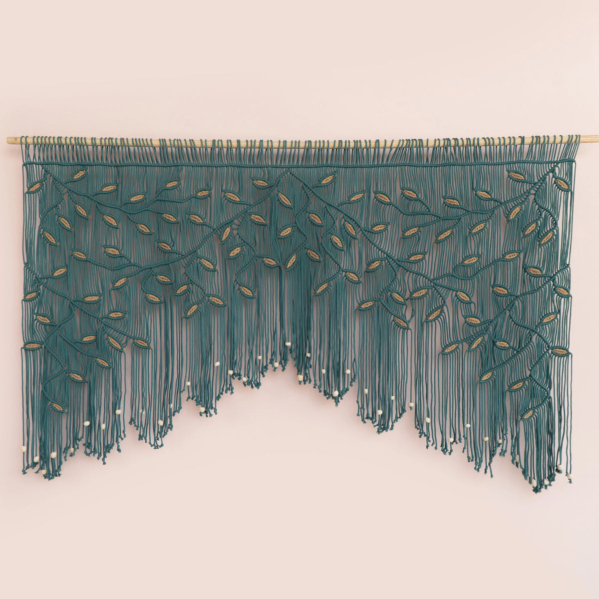 A teal green fringe wall hanging with embroidered leafy branches and small white bead accents, suspended from a light wood rod against a pale pink background.