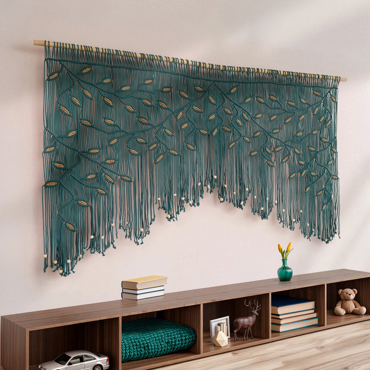A large teal macrame wall hanging with leafy vine patterns and long fringe, displayed on a wall above a wooden storage shelf in a styled room.