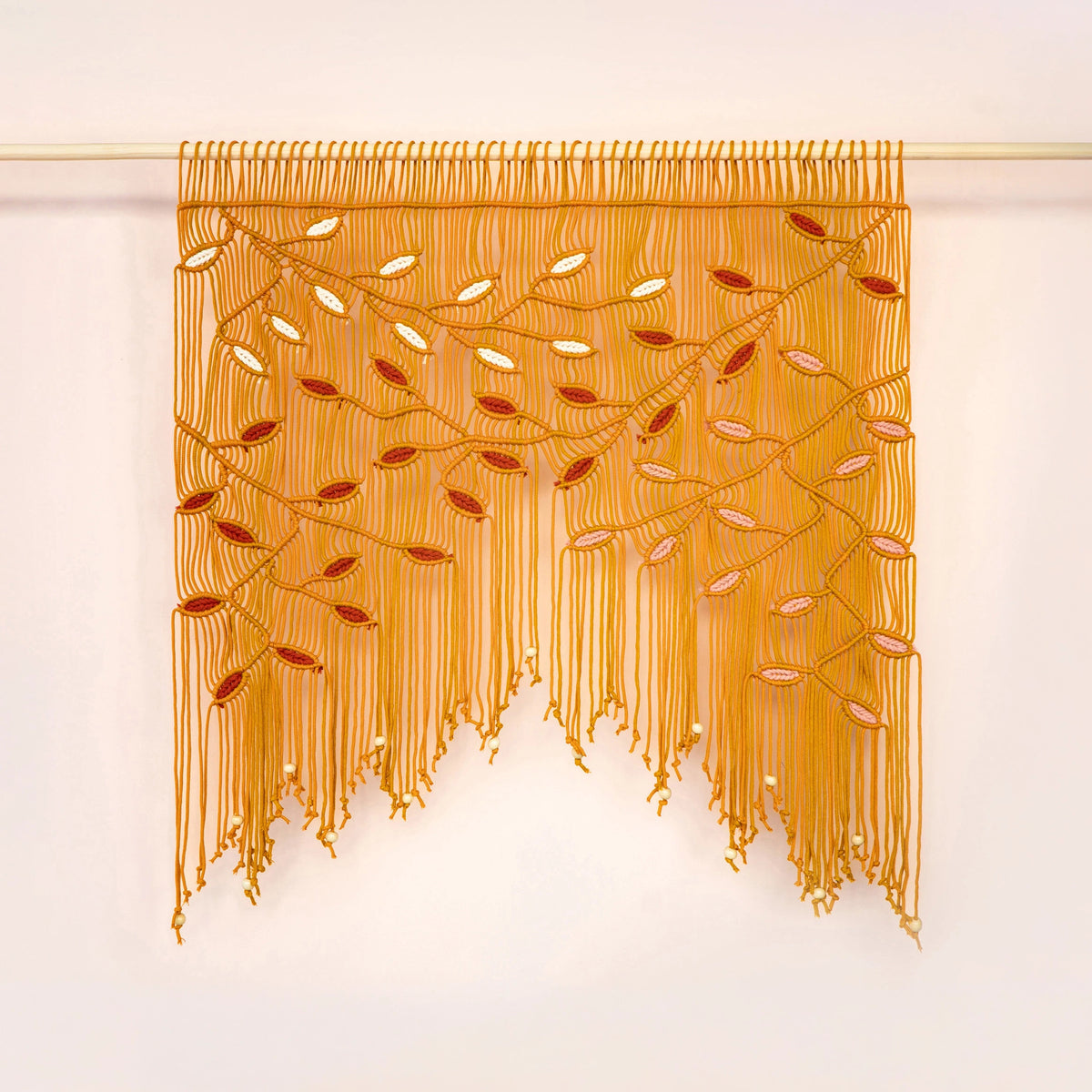 A golden yellow beaded fringe wall hanging with embroidered branch and leaf patterns, suspended from a light wooden rod against a plain off-white wall.