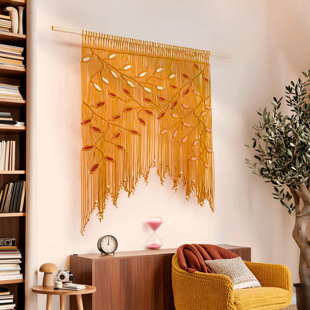 A golden yellow bohemian macrame wall hanging with leaf patterns is displayed on a light beige wall in a styled living room setting with a wooden bookshelf, yellow armchair, and potted plant.