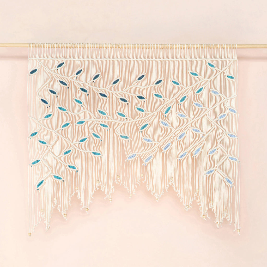 A cream macrame wall hanging with intricate woven leaf patterns and long fringe, suspended from a thin gold rod against a soft pink background.