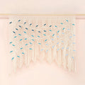 A cream macrame wall hanging with intricate woven leaf patterns and long fringe, suspended from a thin gold rod against a soft pink background.