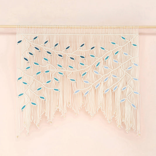 A cream macrame wall hanging with intricate woven leaf patterns and long fringe, suspended from a thin gold rod against a soft pink background.