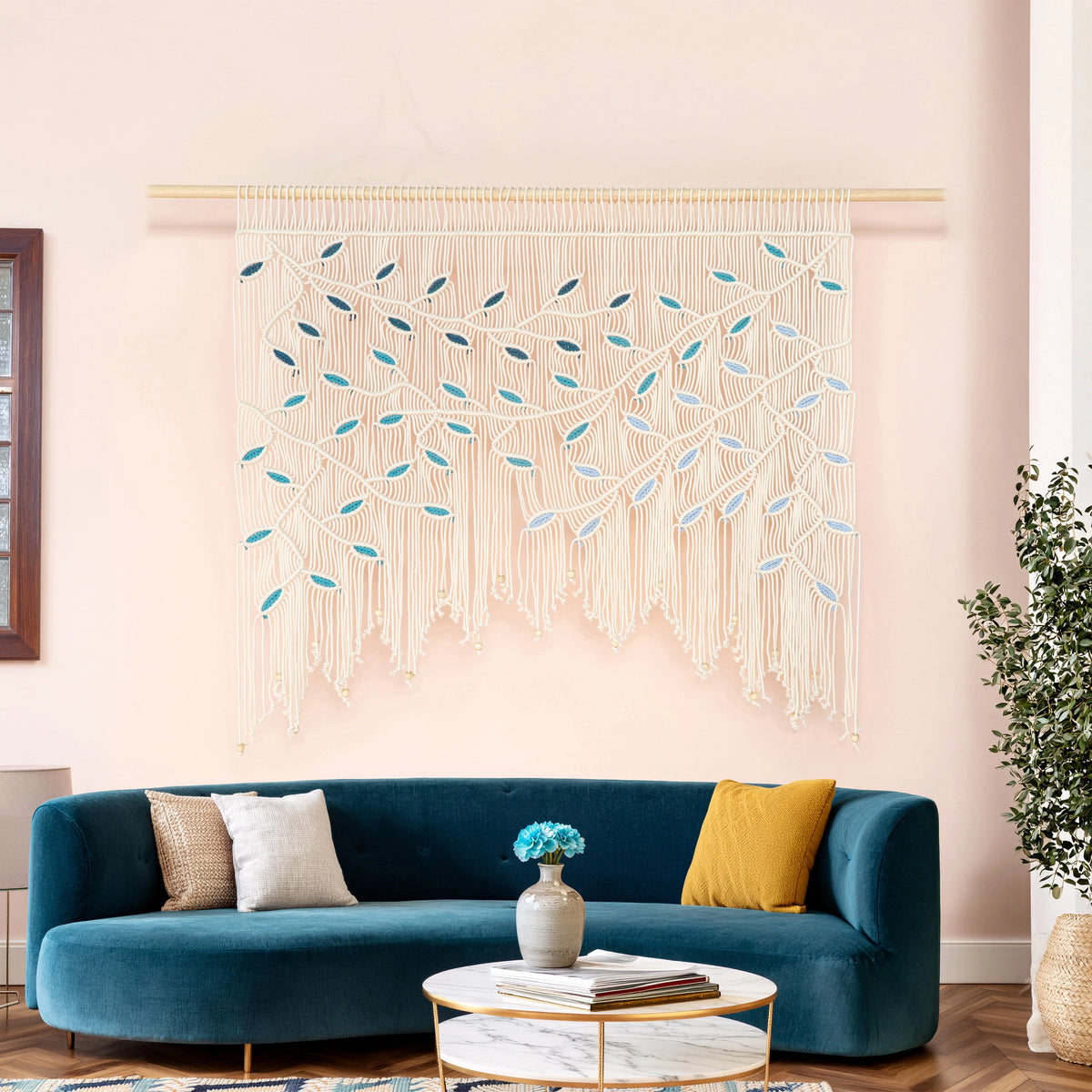 A large bohemian-style macrame wall hanging with a tree branch and leaf pattern in cream, light blue, and teal, displayed on a pink wall above a blue velvet sofa in a styled living room.