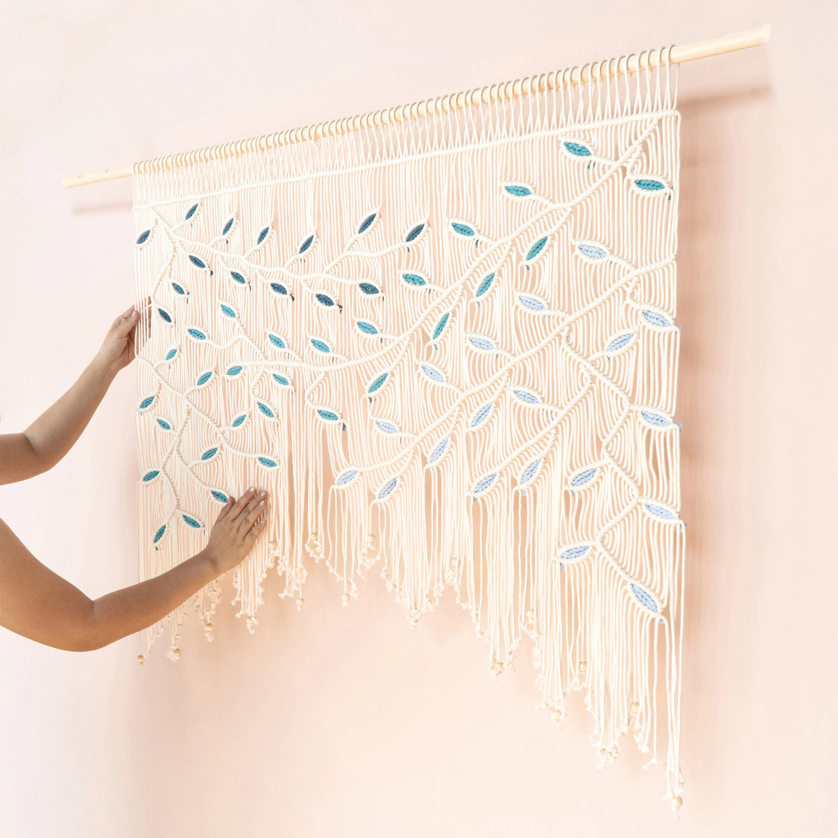 A cream macrame wall hanging with leaf patterns in light blue and white, suspended from a wooden rod on a pale pink wall. A person's hands are visible adjusting the left side of the hanging.