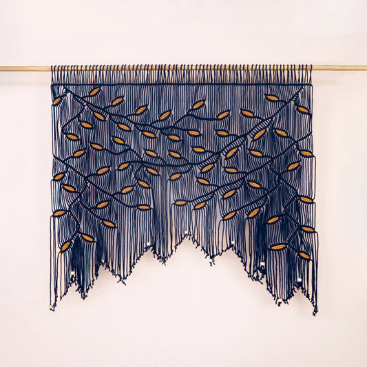 A bohemian-style wall hanging macrame wall tapestry with a tree branch pattern, featuring dark navy blue cords and small gold leaf accents, suspended from a light wood rod against a plain off-white wall.