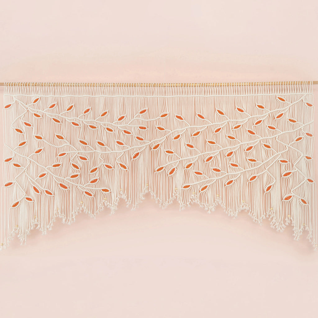 Handcrafted natural beige macrame wall hanging featuring an intricate botanical canopy of cream cords and vibrant orange leaves, hung above a rust-colored velvet sofa.