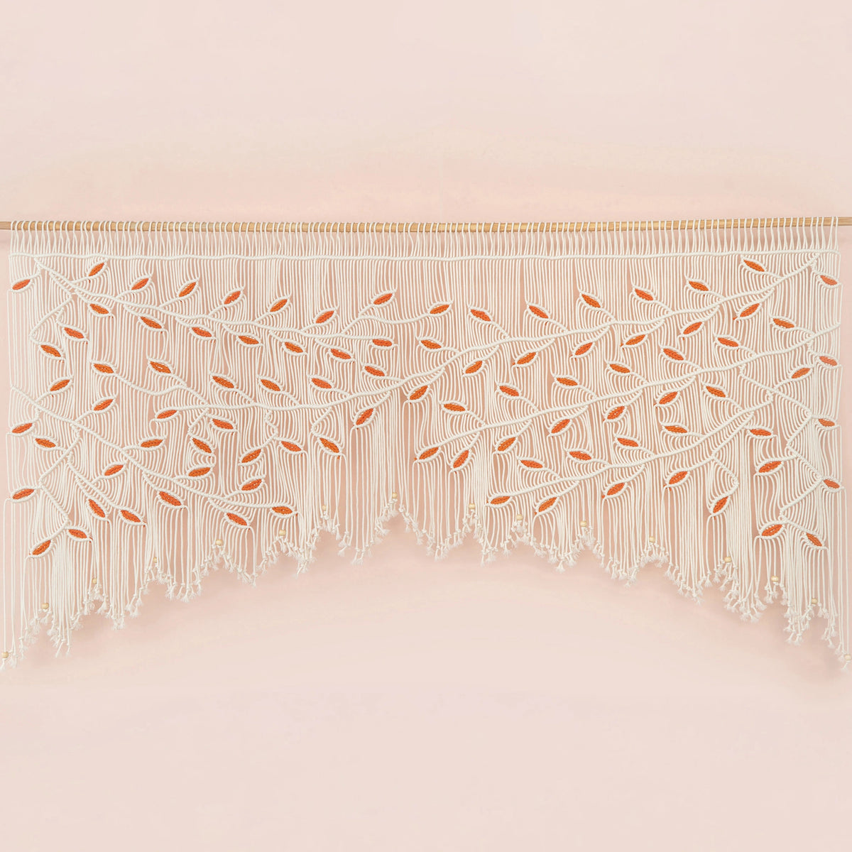Handcrafted natural beige macrame wall hanging featuring an intricate botanical canopy of cream cords and vibrant orange leaves, hung above a rust-colored velvet sofa.