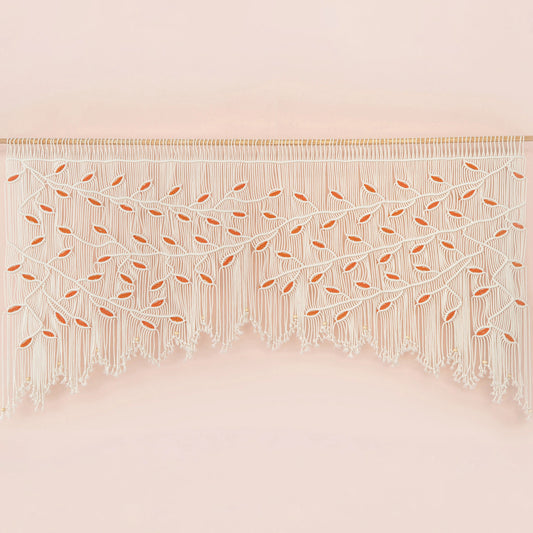 Handcrafted natural beige macrame wall hanging featuring an intricate botanical canopy of cream cords and vibrant orange leaves, hung above a rust-colored velvet sofa.