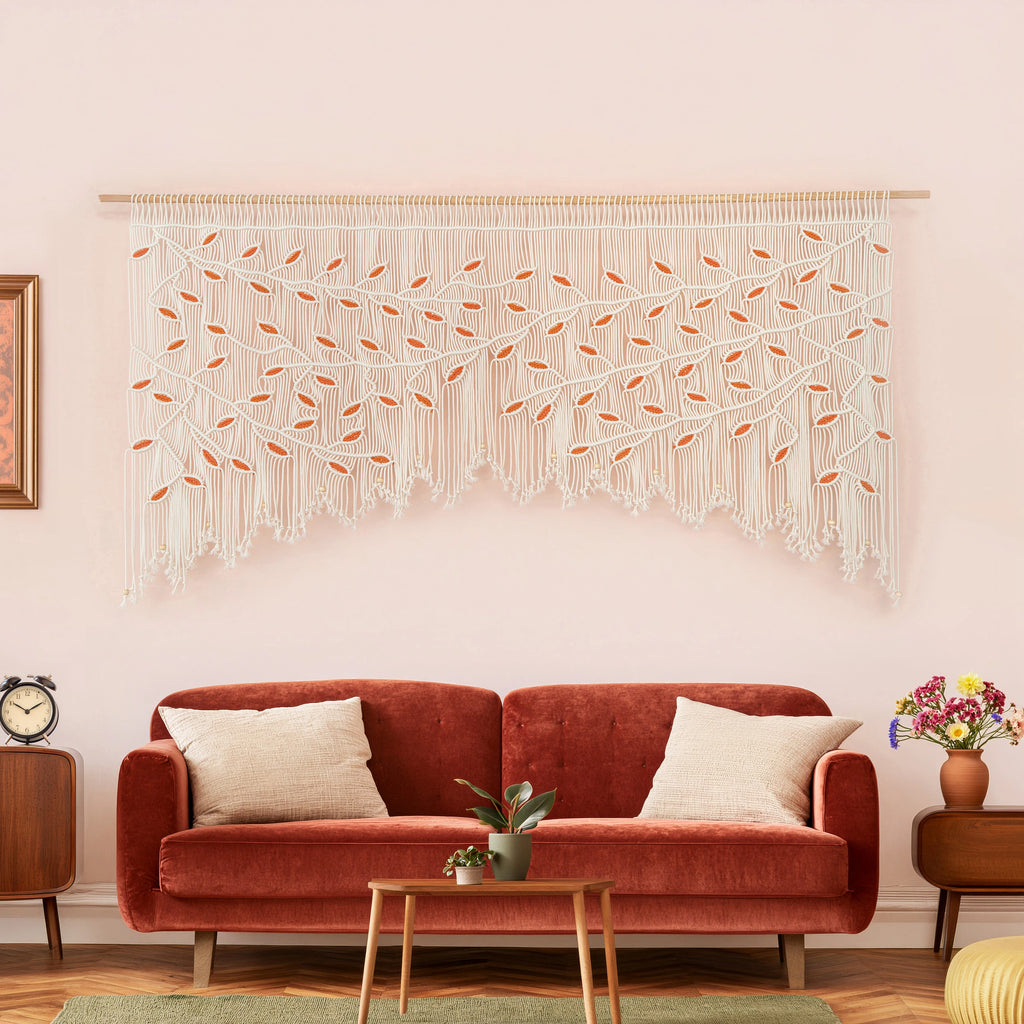 Large natural and orange leaf macrame wall art hanging horizontally on a smooth wooden dowel against a soft pinkish-neutral interior wall.