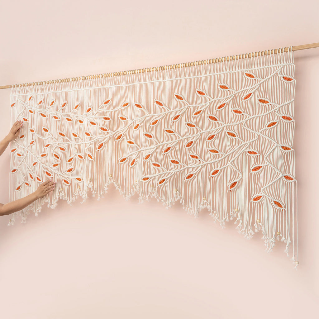 Detailed close-up of a creamy natural macrame wall hanging showing precise knotwork and contrasting vibrant orange botanical leaf motifs over a modern seating area.