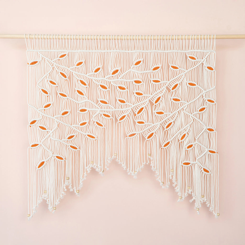 Handcrafted natural beige macrame wall hanging featuring an intricate botanical canopy of cream-colored vines and vibrant orange leaves.