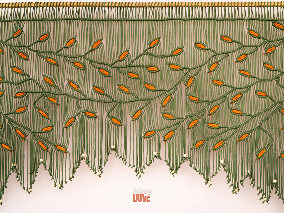 Large olive green and orange leaf macrame wall art hanging horizontally on a smooth wooden dowel against a soft pinkish-neutral interior wall.