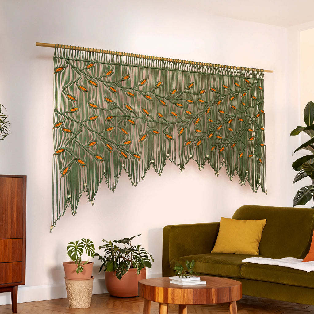 Wide botanical macrame tapestry showcasing an arched branch canopy in dark green with warm rust-orange leaf highlights positioned above a velvet sofa.