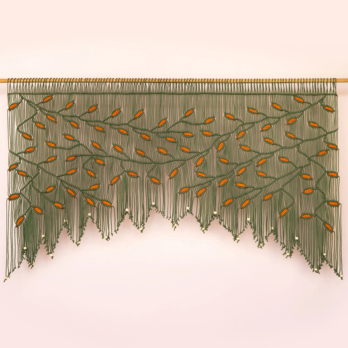 A decorative wall hanging or curtain made of many long green strands, arranged in a tree-like pattern with small orange leaf shapes and a gold rod across the top, shown against a pale pink background.