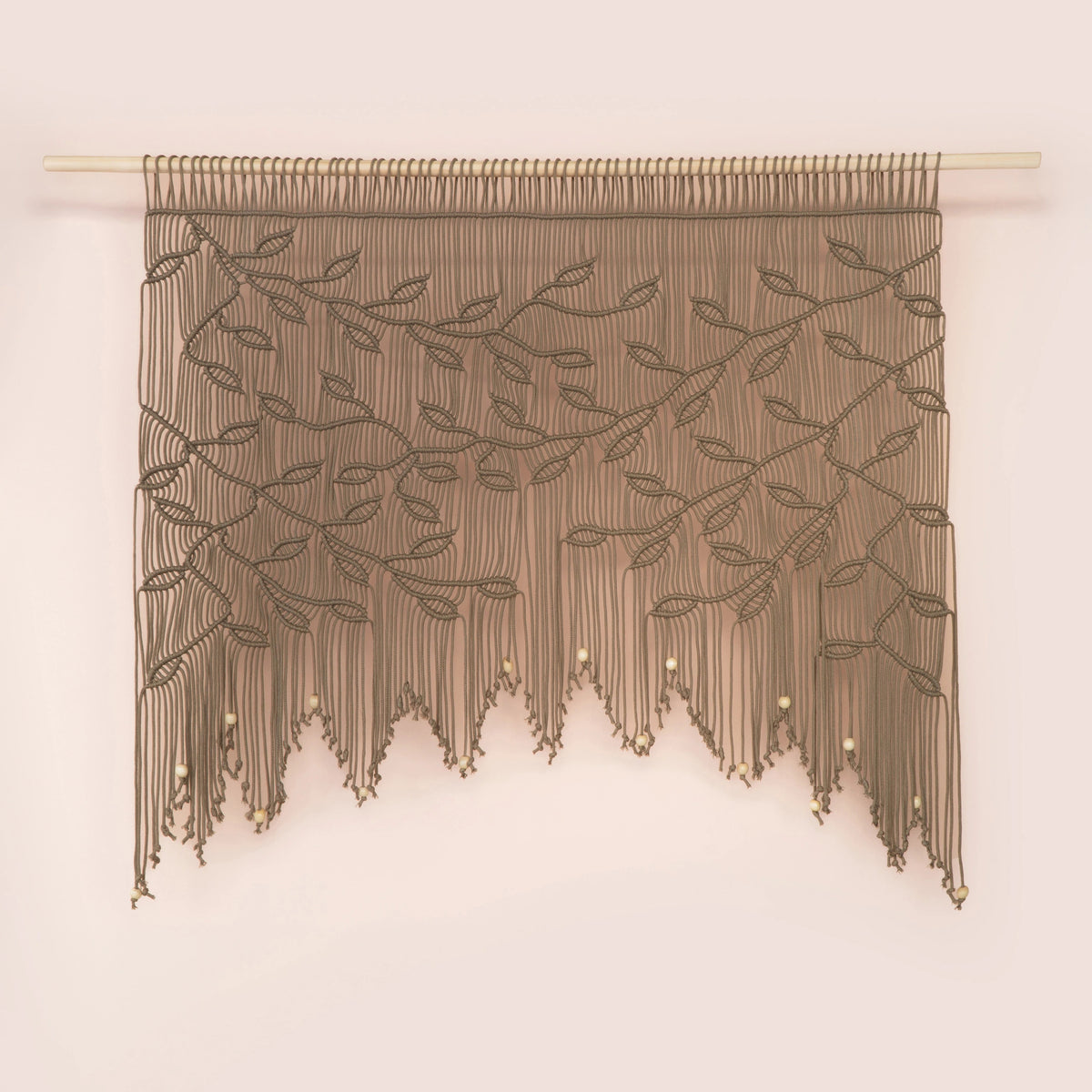 A brown macrame wall hanging with a leafy vine pattern and dangling fringe, suspended from a light wooden dowel against a pale pink wall.