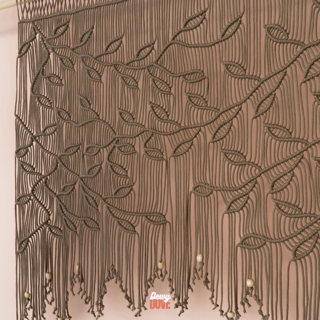 A close-up of a brown macrame wall hanging with leafy vine patterns and hanging tassels, shown against a plain light background. A small 'Davy Cute' watermark is visible near the bottom.