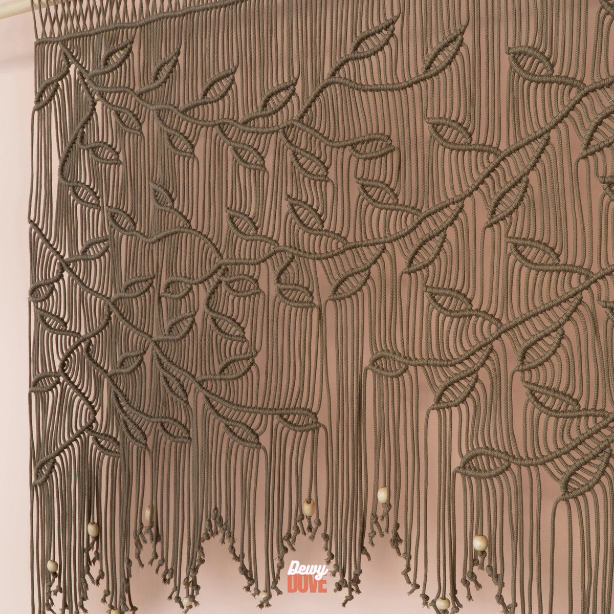 A close-up of a brown macrame wall hanging with leafy vine patterns and hanging tassels, shown against a plain light background. A small 'Davy Cute' watermark is visible near the bottom.