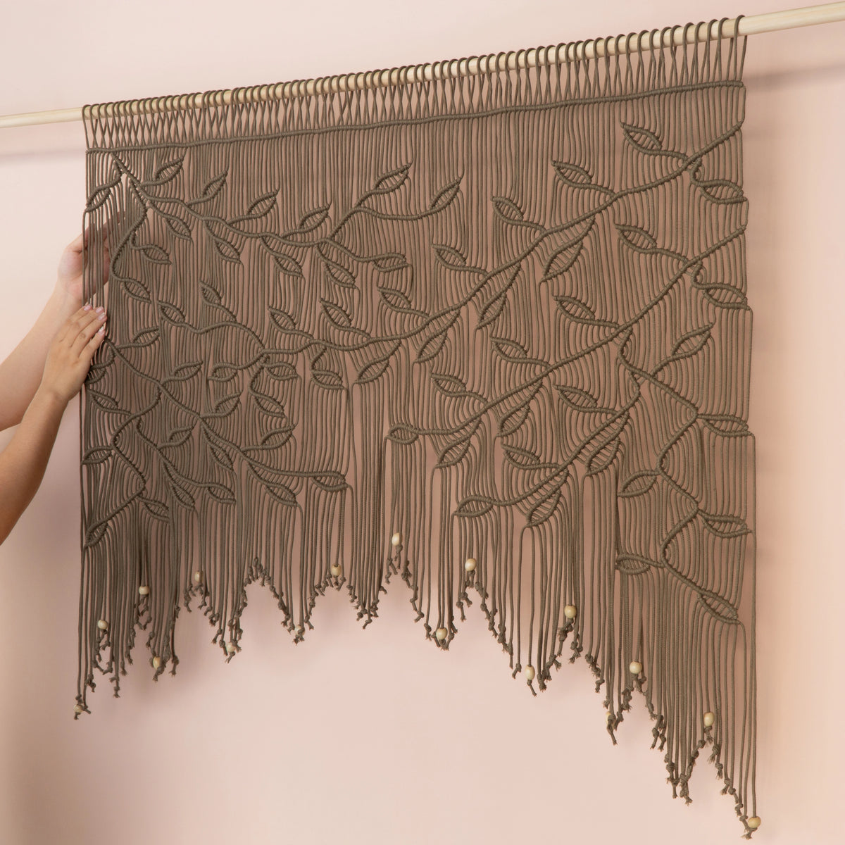 A large woven wall hanging with an intricate leaf and vine pattern, suspended from a rod against a pale pink wall. A person's hands are visible on the left side holding the fabric in place.