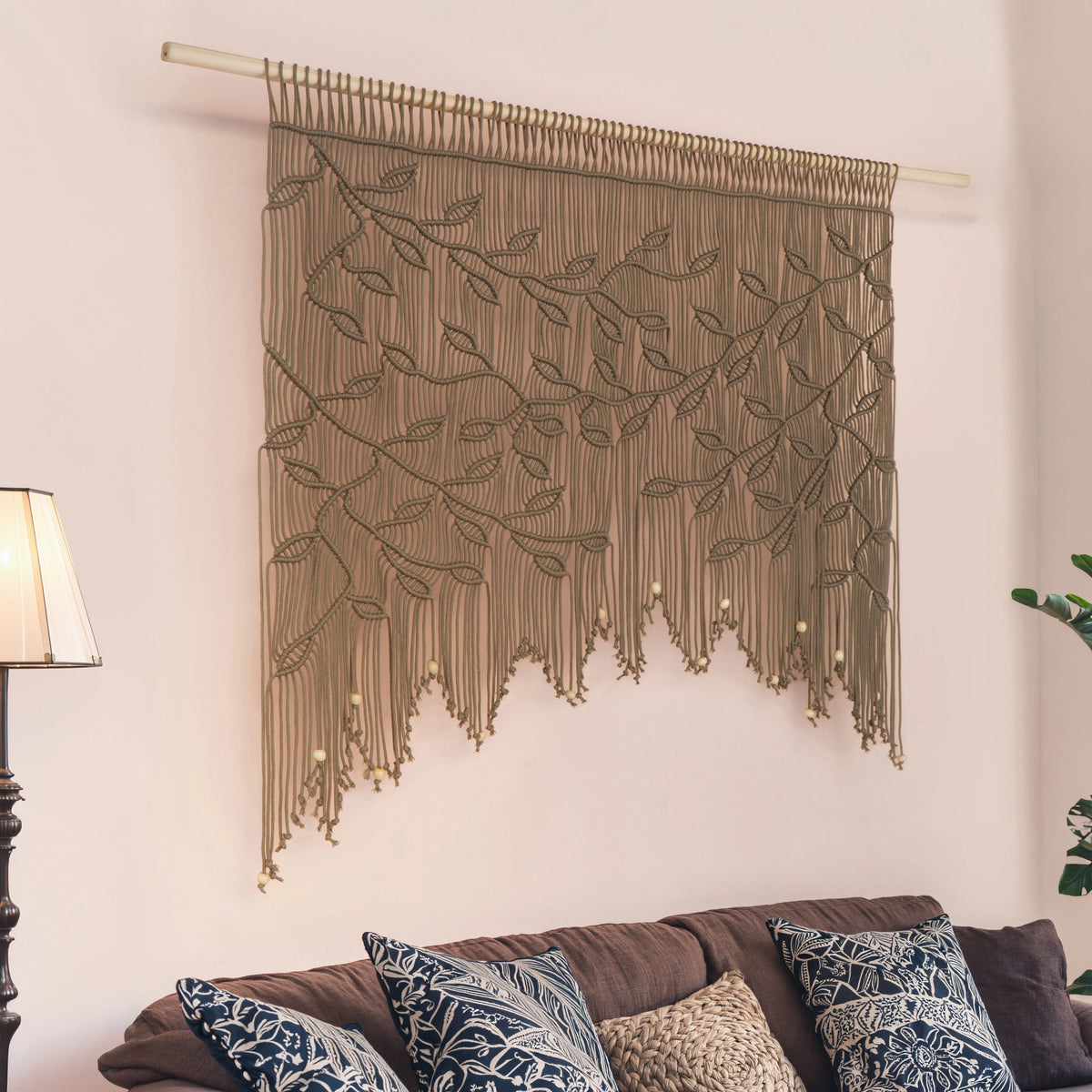 A large woven wall hanging with an embroidered leafy branch pattern and long fringe, suspended from a light wood rod above a sofa in a living room setting.