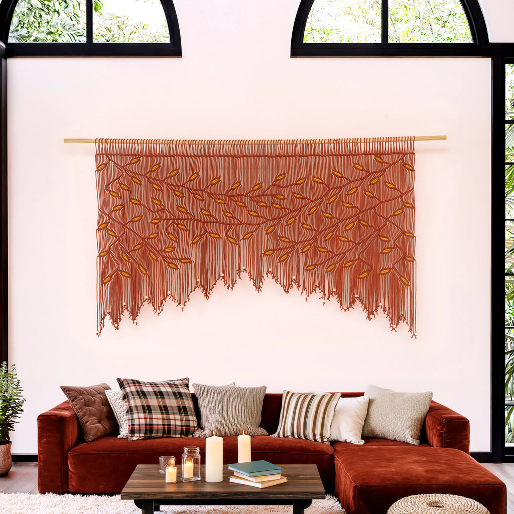 A large bohemian-style woven wall tapestry with a tree branch and leaf pattern in terracotta and mustard yellow, hanging on a light wood rod above a rust-brown sectional sofa in a modern living room setting.