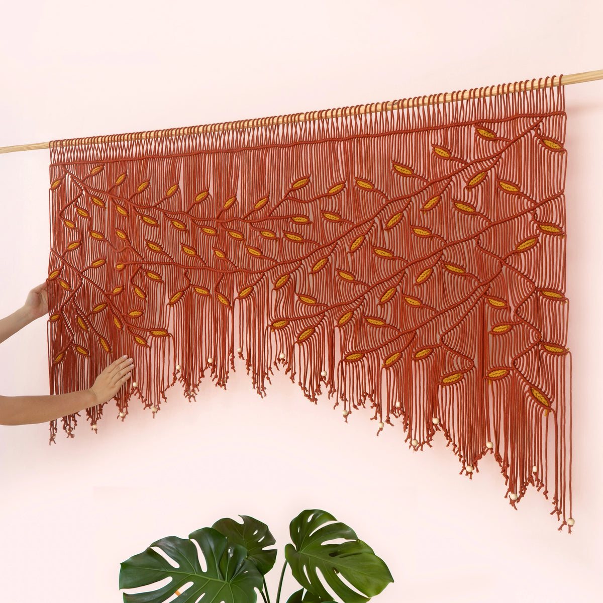 Handcrafted terracotta macrame wall hanging featuring an intricate botanical canopy of burnt orange vines and golden mustard yellow leaves.