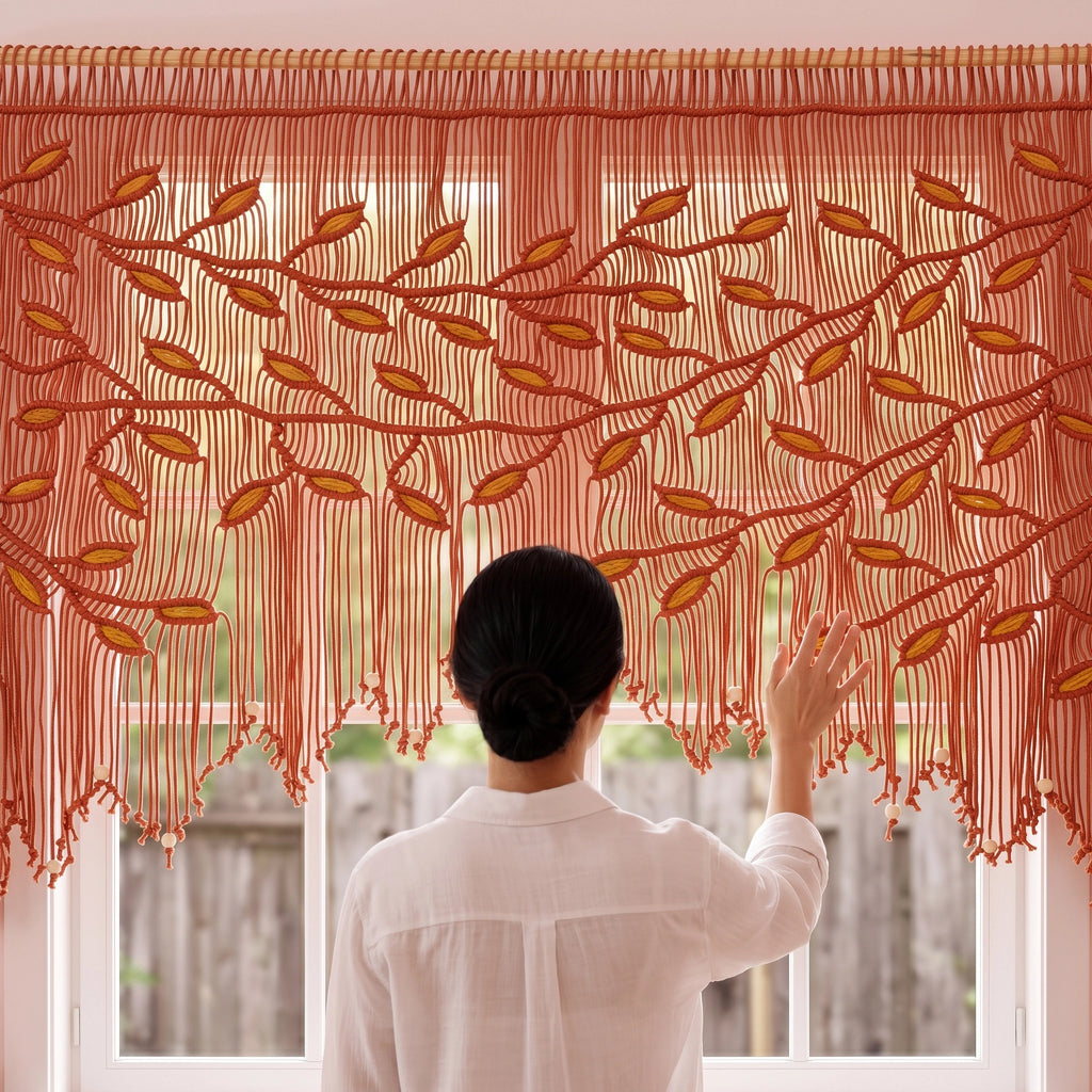 A woman viewed from behind stands in front of a window wearing a sheer rust-orange valance with embroidered leafy branches and fringe, touching the fabric.