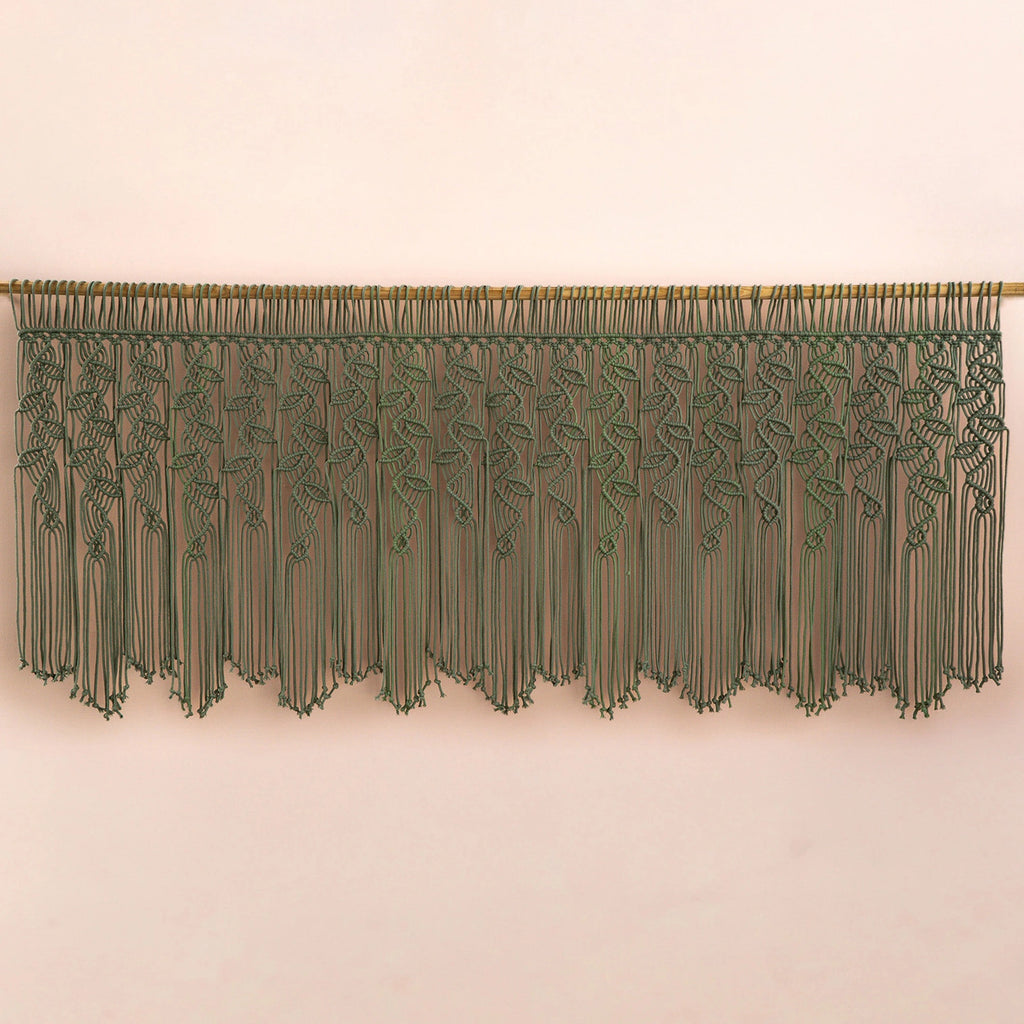 An olive green macrame wall hanging with a woven vine pattern and long fringe, suspended from a wooden rod against a plain beige wall.