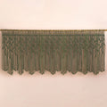 An olive green macrame wall hanging with a woven vine pattern and long fringe, suspended from a wooden rod against a plain beige wall.