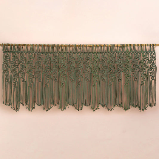 An olive green macrame wall hanging with a woven vine pattern and long fringe, suspended from a wooden rod against a plain beige wall.