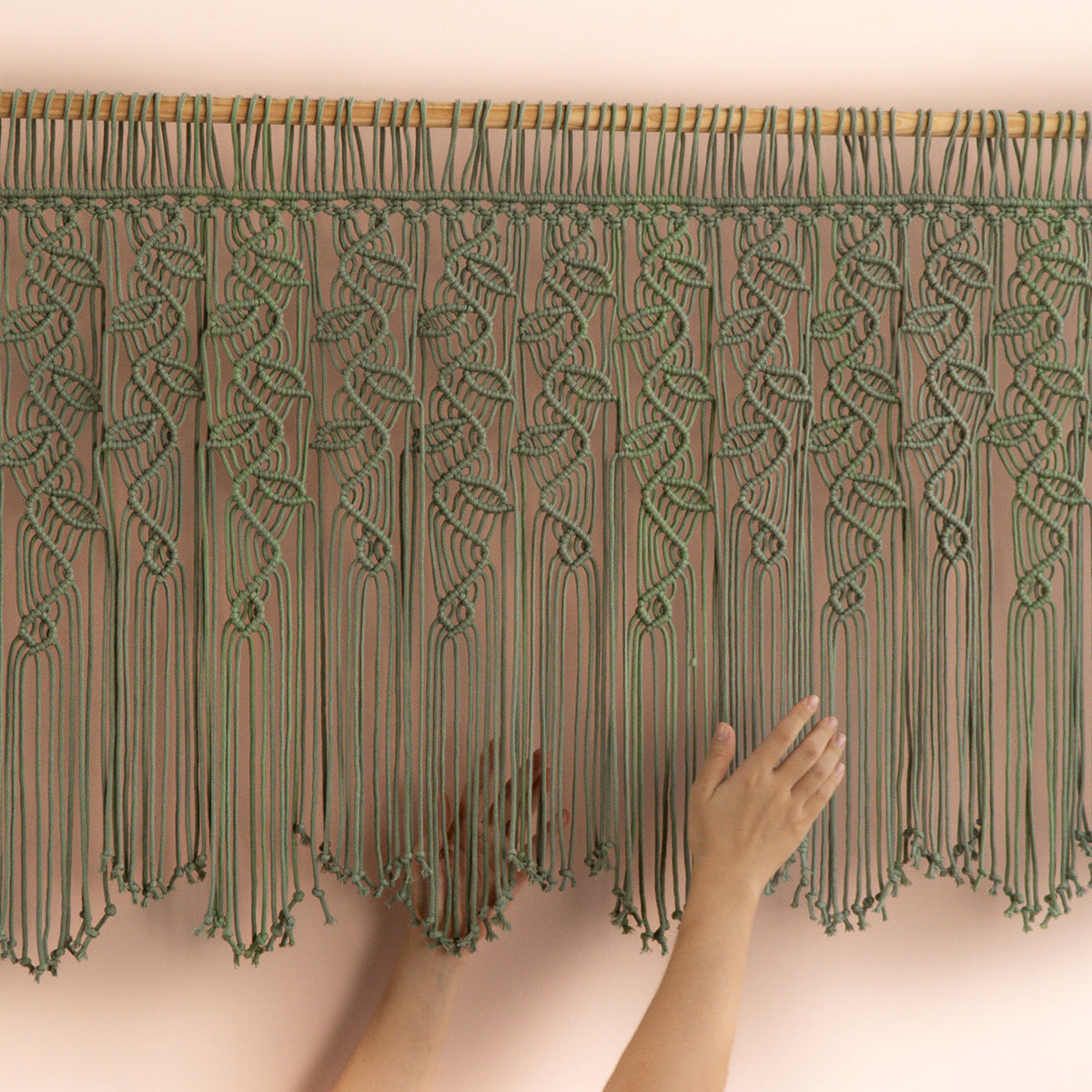 Handmade macrame wall hanging with a hand touching it against a plain background